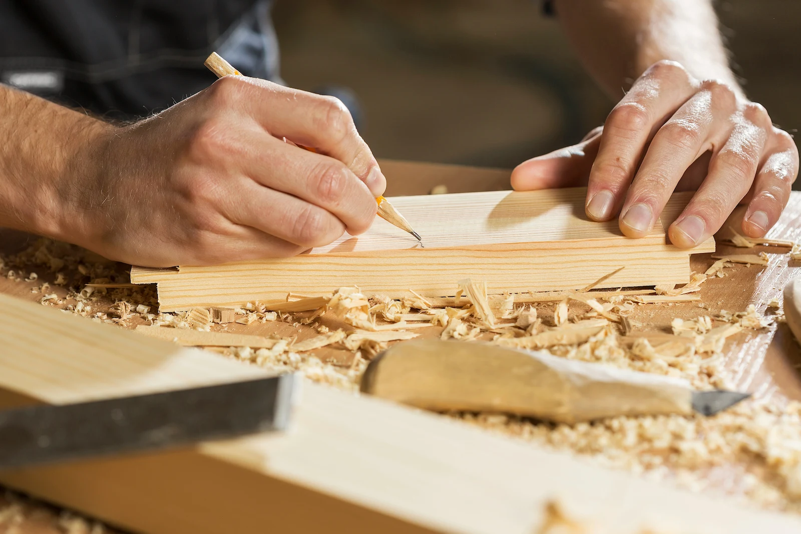 Close-up of hands marking a measured line on a wood plank with a pencil, wood shavings and a chisel scattered across the workbench